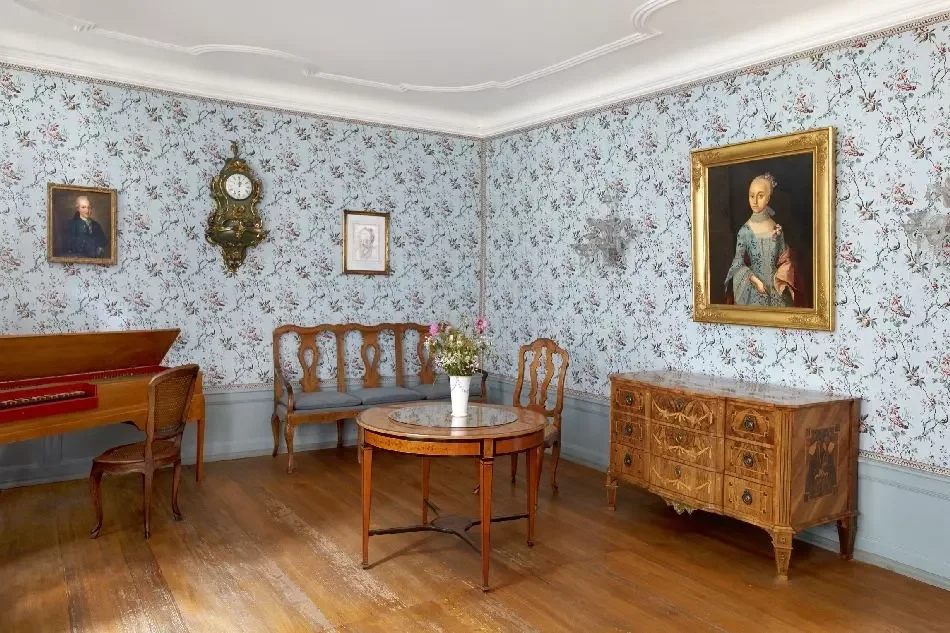 Zimmer mit antiken Möbeln, blau-geblümter Tapete und Gemälden und Uhr an der Wand